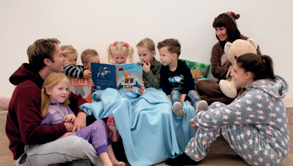 Groep kleuters wordt voorgelezen uit prentenboek, zit aandachtig om het boek heen an kijkt en wijst naar de plaatjes.