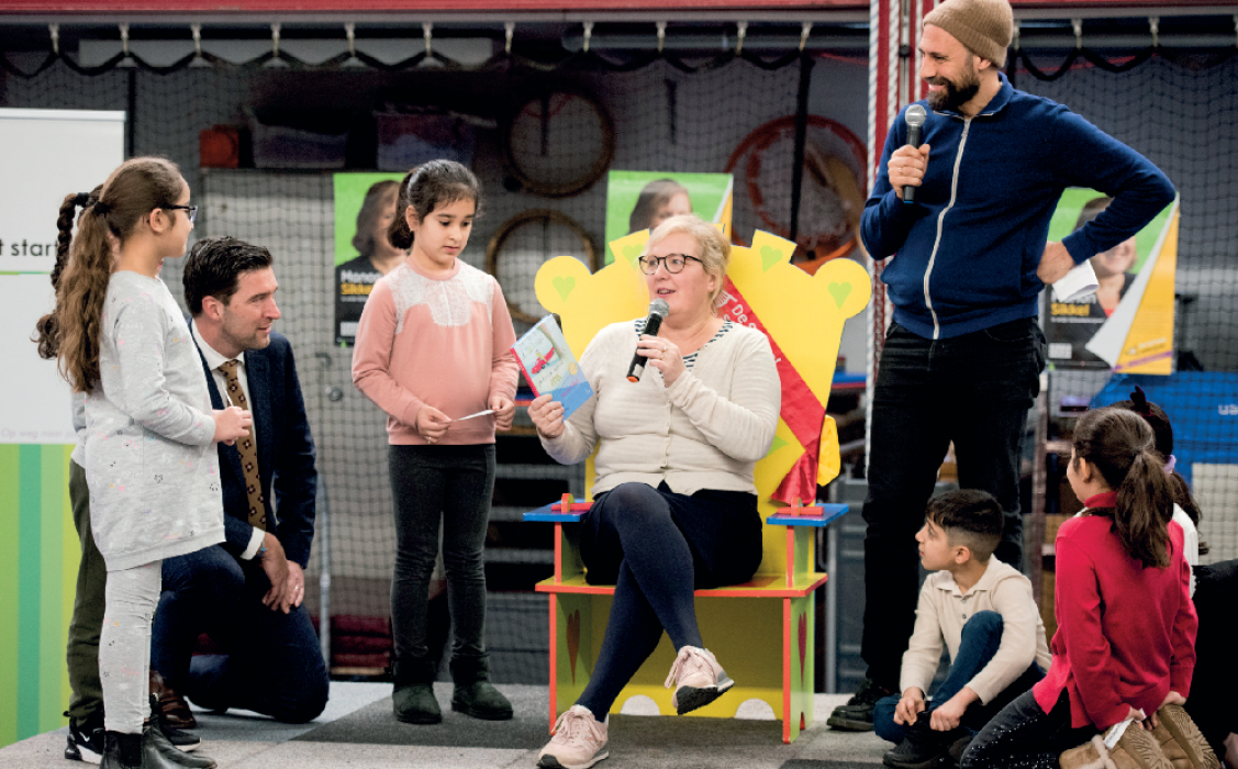 Kinderboekenambassadeur zit op stoel en leest voor, rechts vertelt Abdelkader Benali. Kinderen luisteren aandachtig. 