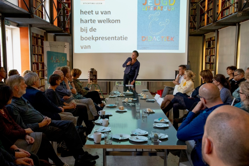 Tafel met aanwezigen eromheen. Gerlien van Dalen start presentatie aan het hoofd van de tafel, met op de achtergrond een powerpointpresentatie. 