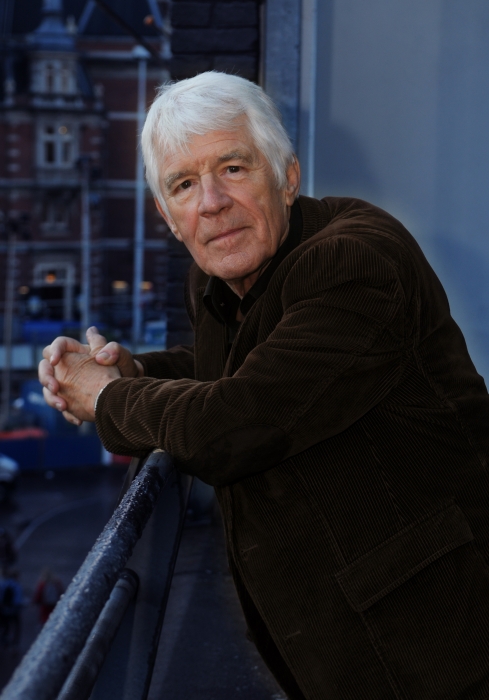 Portretfoto Kees van Kooten, midden in beeld, hij kijkt recht in de camera en leunt op de balustrade van een balkon met ineengestrengelde handen.