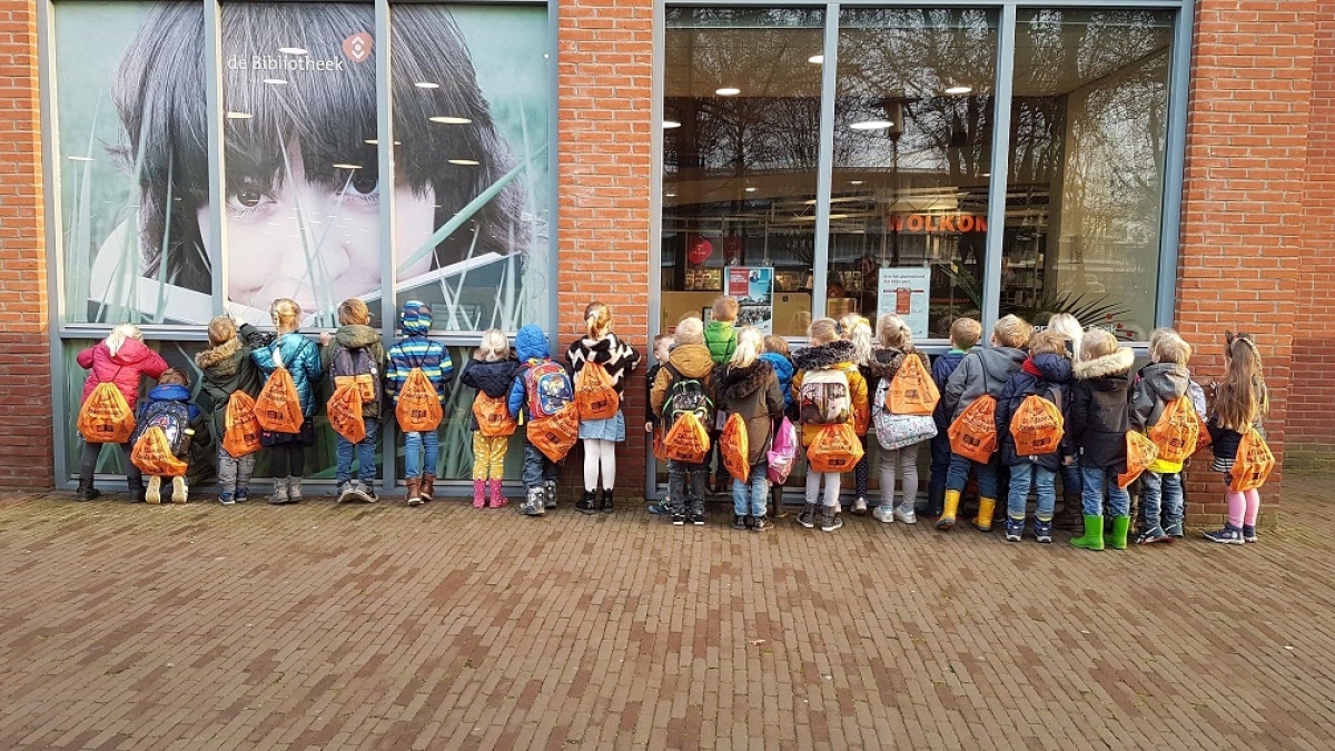 Kinderen met voorleestas voor de bieb in Heerenveen. Kinderen staan met rug naar de camera toe, voorleestassen zijn op deze manier goed in beeld. 