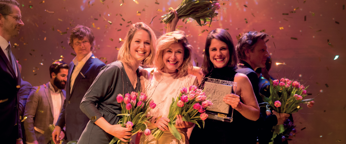 Schrijverscollectief Schik (midden) winnen dePublieksprijs,. Van links naar rechts hebben twee dames tulpen vast. De derde heeft de prijs, een tegel, vast. Confetti dwarrelt door het beeld.