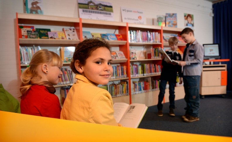 Twee meisjes op een gele bank in de schoolbibliotheek, op de achtergrond staan twee jongens bij de boekenkast