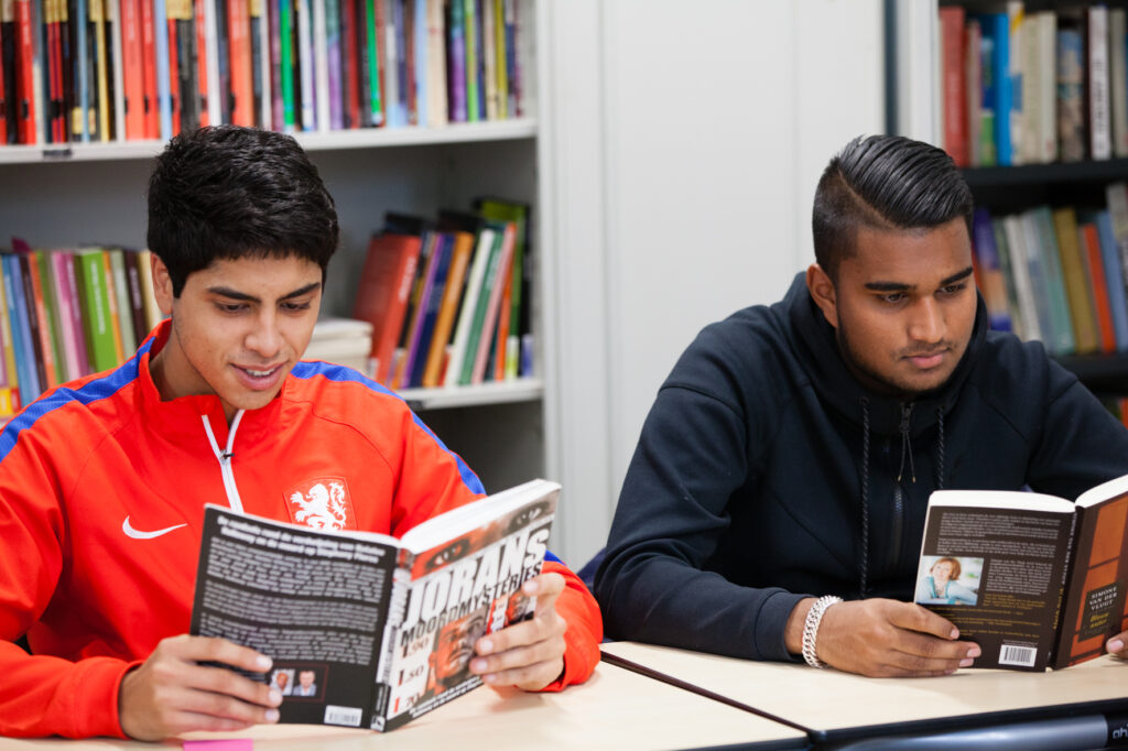 Twee mbo-studenten lezend aan hun tafeltjes in een lokaal met boekenkast op de achtergrond