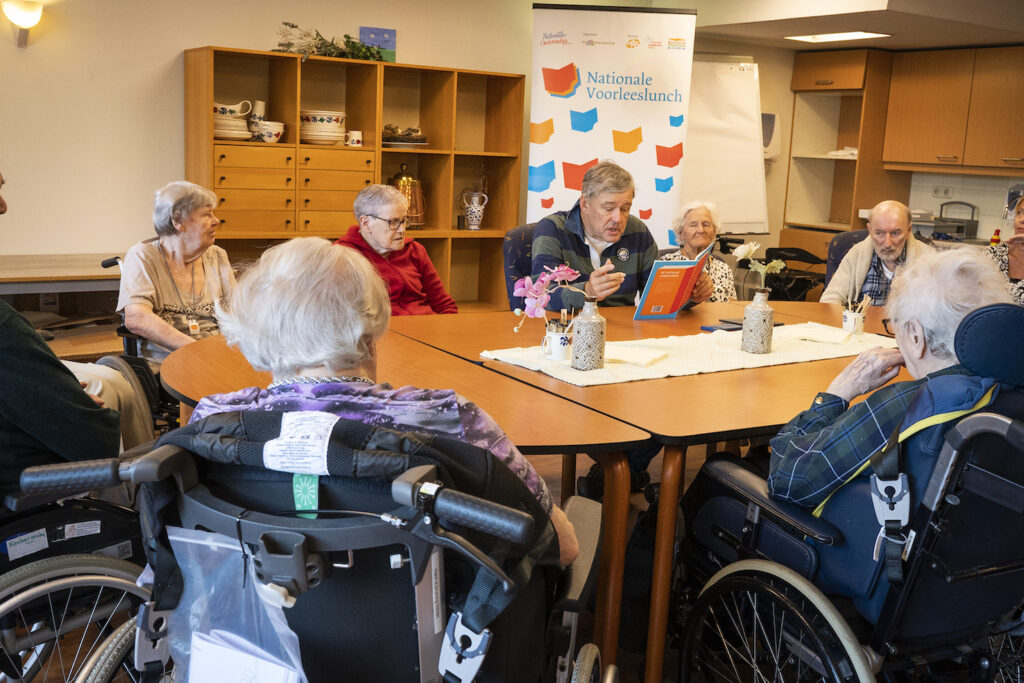 Charles Groenhuijsen (midden) leest voor aan tafel met ouderen.