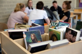 Boeken op de voorgrond, op de achtergrond zijn leerlingen met elkaar in gesprek in een klaslokaal. 