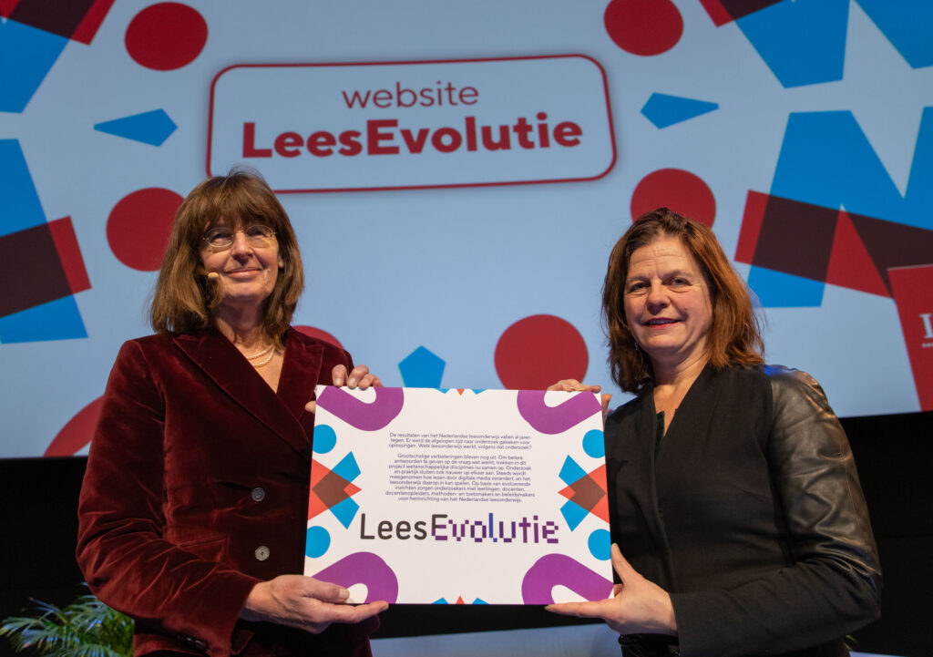 Els Stronks (links) en Inge Vossenaar houden het bord met de startverklaring van het onderzoek LeesEvolutie vast, met op de achtergrond een PowerPoint met knop website LeesEvolutie.