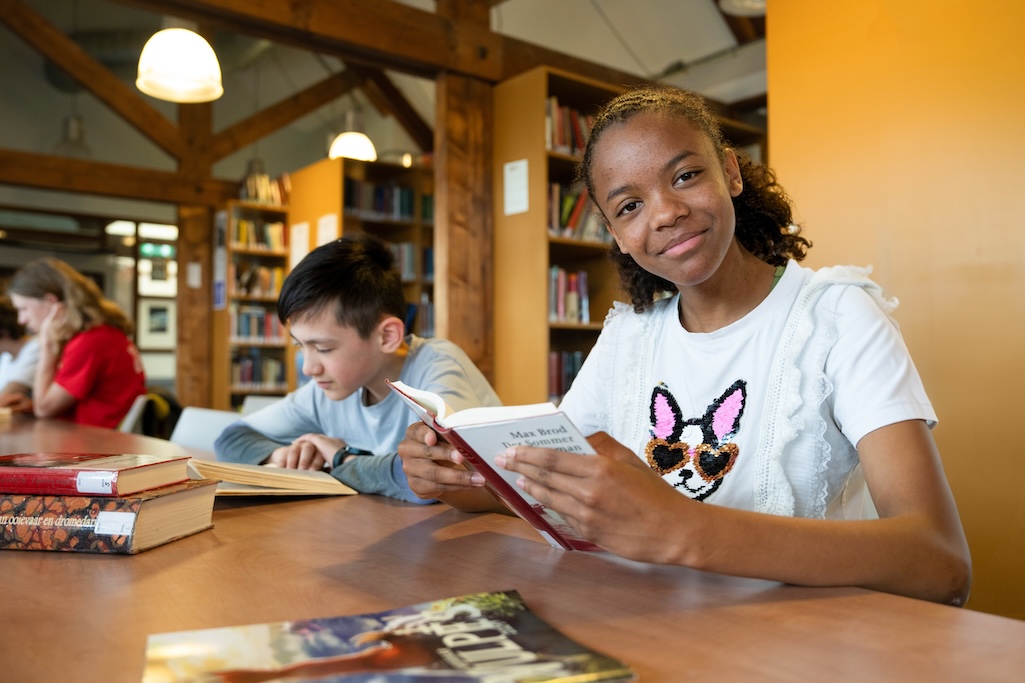 Leerling zit aan tafel in een mediatheek en leest een boek. De leerling kijkt in de camera.