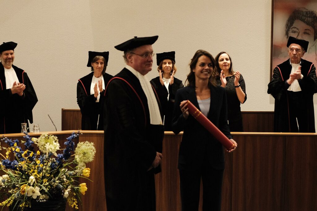 Linda (rechts) ontvangt haar doctorsbul van haar promotor. Op de achtergrond applaudisseert de commissie.