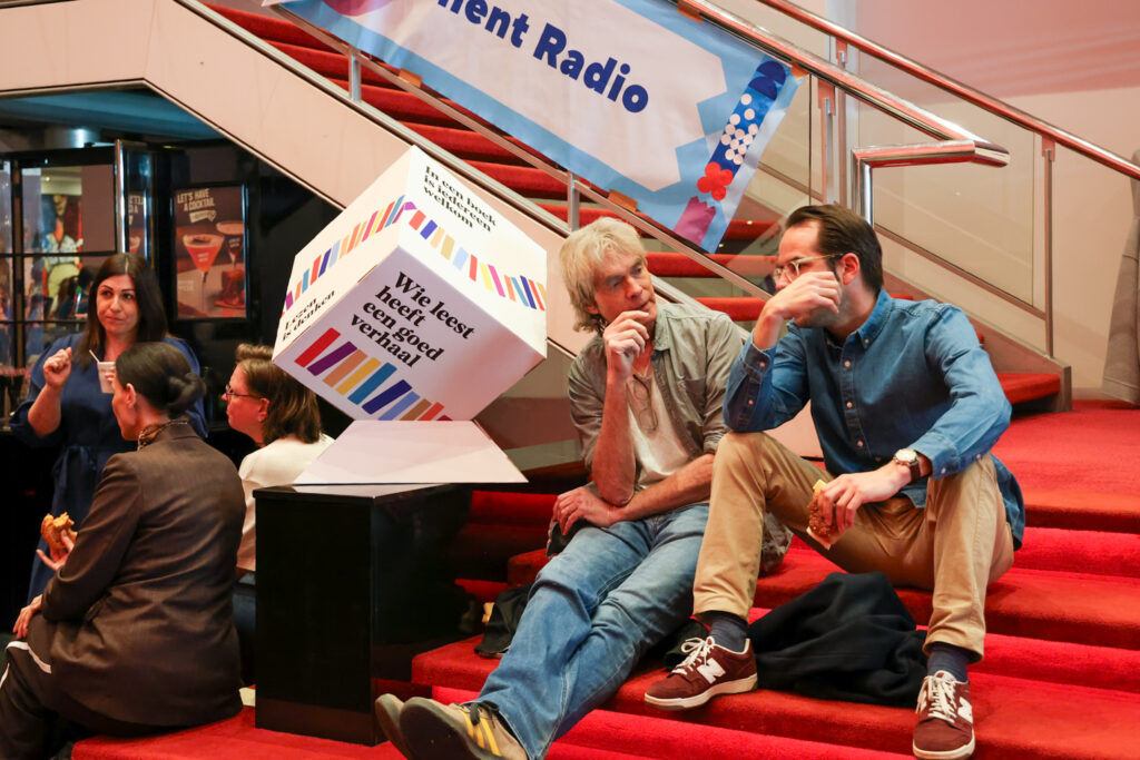 Twee deelnemers aan Lezen Centraal praten met elkaar, zittend op de trap. Op de achtergrond een kubus met 'Wie leest heeft een goed verhaal'